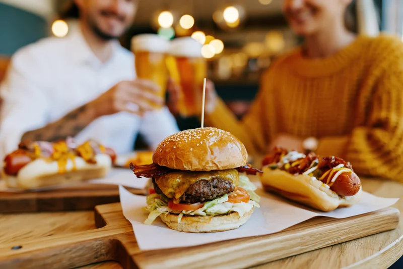Loaded burger on wooden board with hotdogs and beer, couple toasting in background