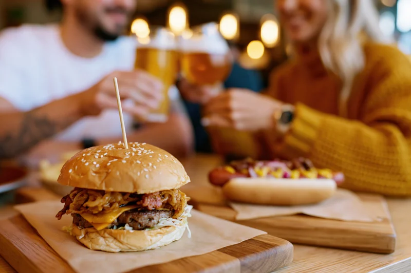 Loaded burger with cheese and bacon on wooden board in foreground with couple toasting beer in background