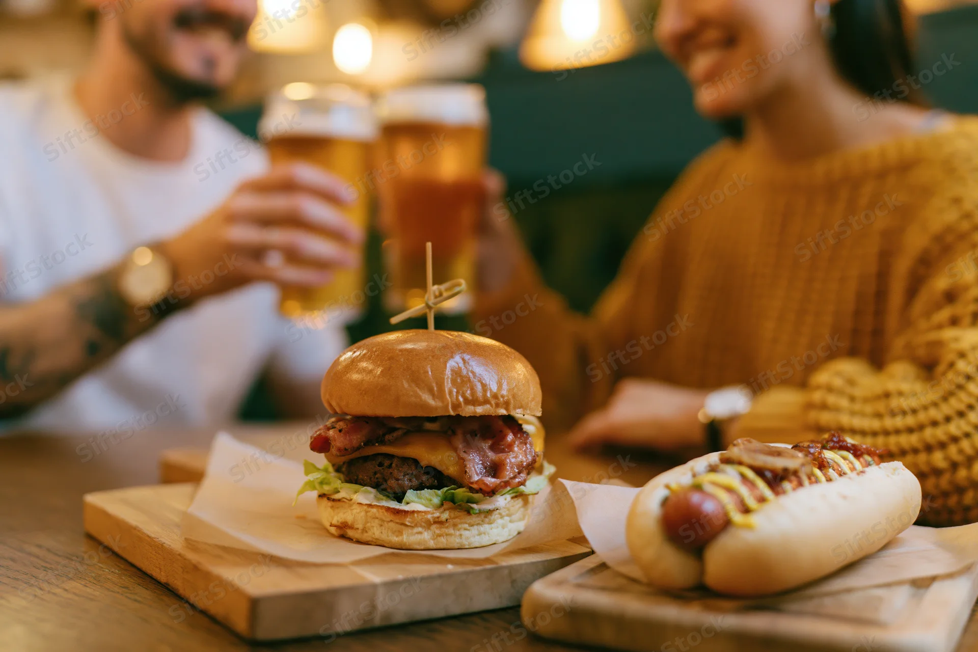 Loaded burger and hotdog on wooden boards with couple toasting beer in background