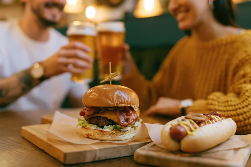 Loaded burger and hotdog on wooden boards with couple toasting beer in background