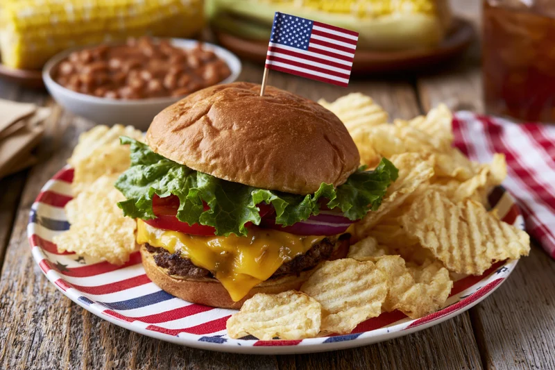 Cheeseburger with lettuce tomato onion and cheese on a patriotic plate with potato chips and american flag toothpick, served with corn and beans on wooden table