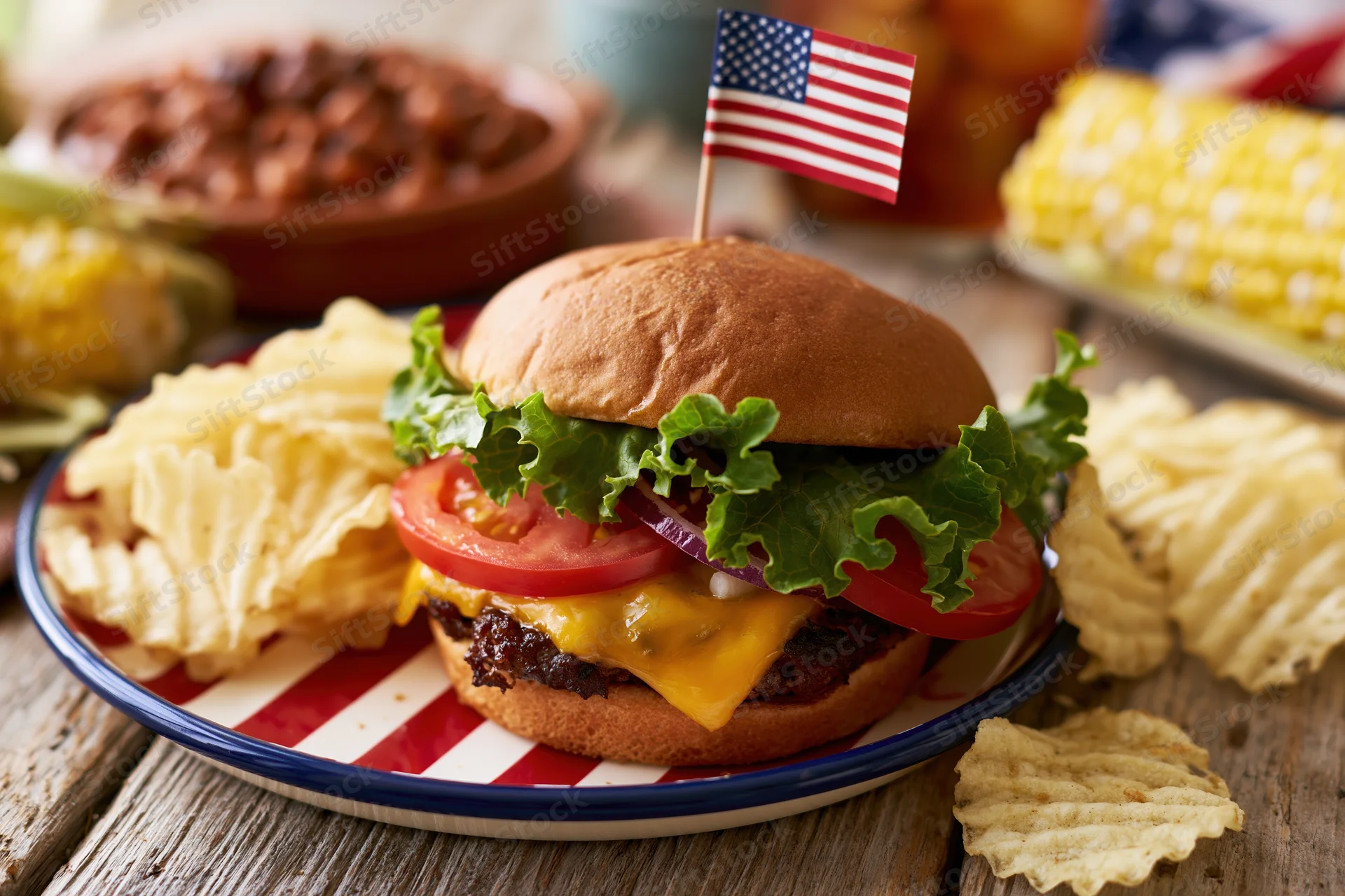Cheeseburger with lettuce tomato and cheese on a red and white striped plate with an american flag toothpick, surrounded by potato chips and corn on a wooden table