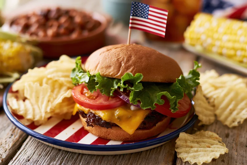 Cheeseburger with lettuce tomato and cheese on a red and white striped plate with an american flag toothpick, surrounded by potato chips and corn on a wooden table