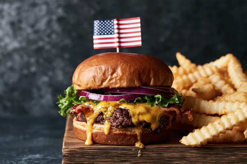 Loaded cheeseburger with melted cheese dripping down and american flag on top served with crinkle fries on wooden board