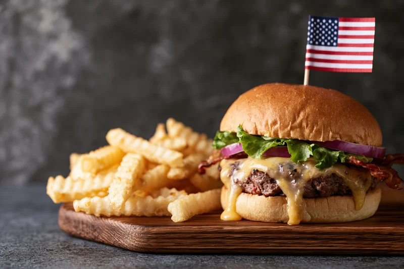 Cheeseburger with melted cheese dripping down and american flag on top served with crinkle fries on wooden board
