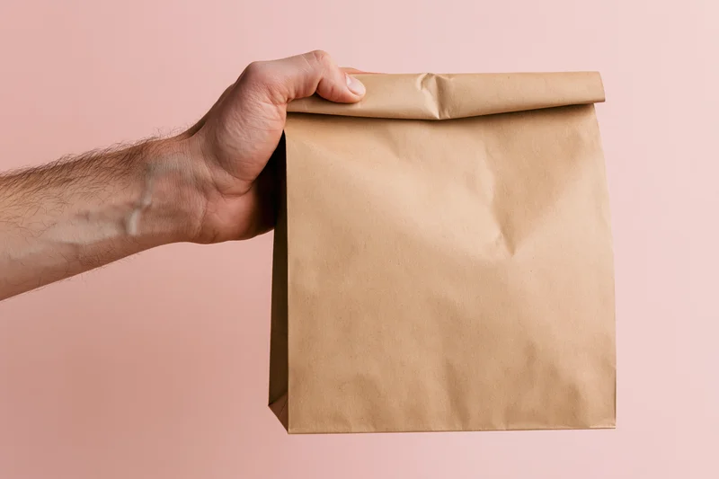 Hand gripping folded top of plain kraft paper bag against pink background