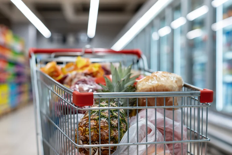 Metal shopping cart filled with groceries including pineapple and bread in supermarket aisle