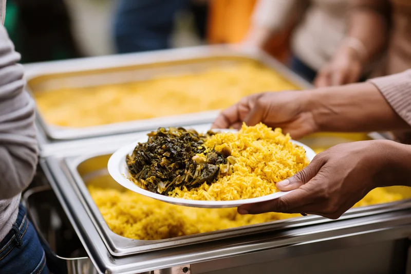 Hands holding a plate with yellow rice and collard greens being served from a buffet tray