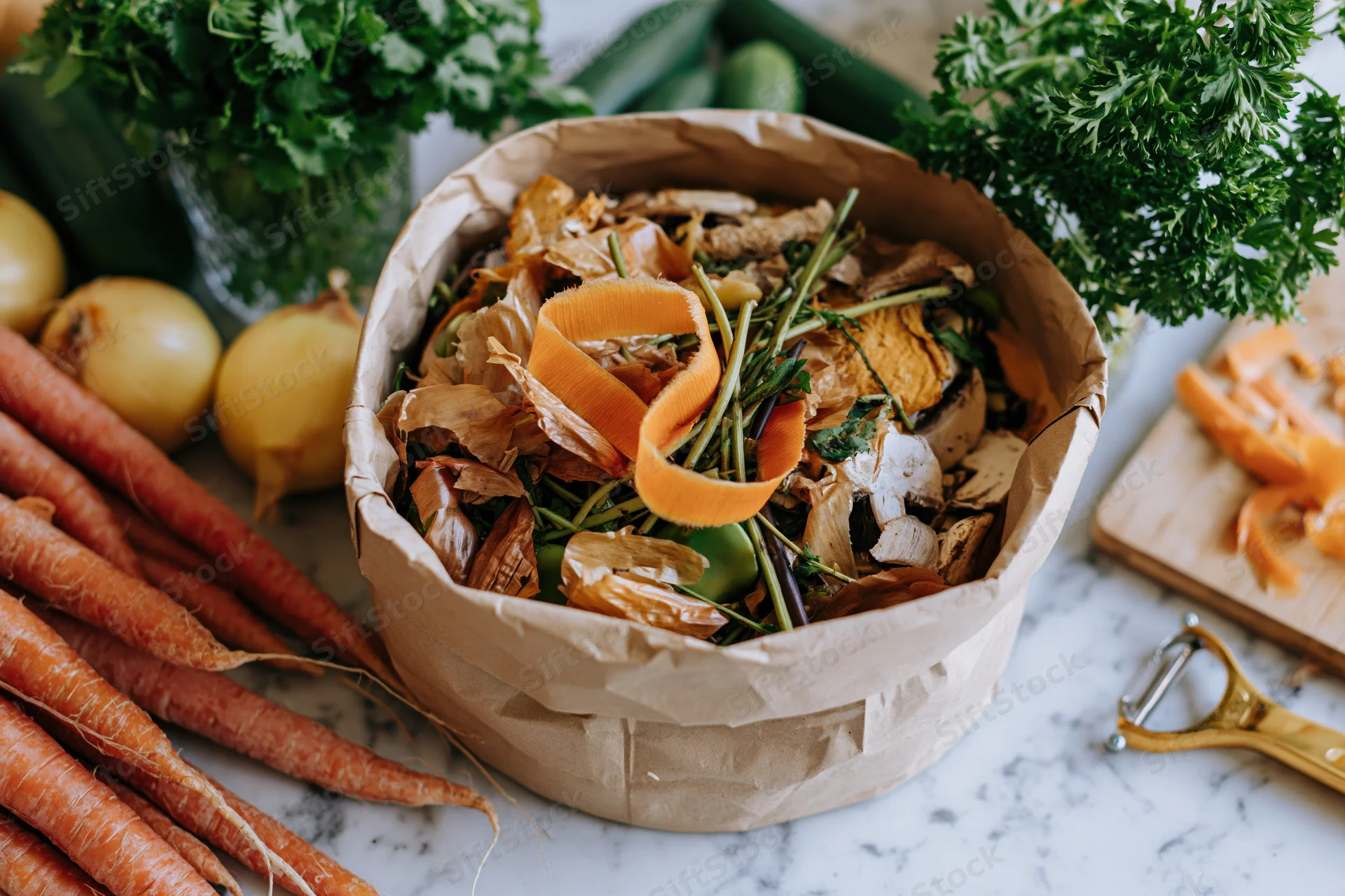 Paper bag filled with vegetable scraps and peels on a marble countertop surrounded by fresh produce and a peeler