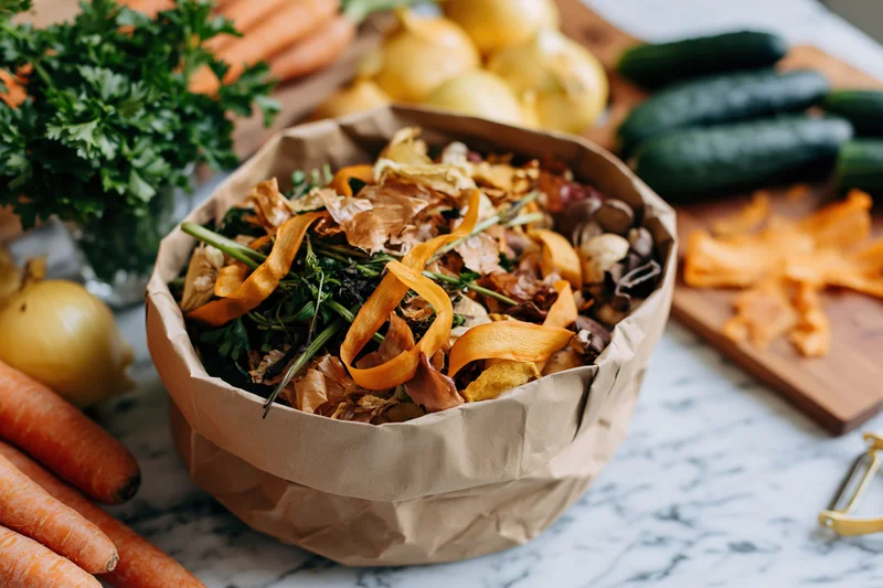 Paper bag filled with vegetable scraps and peels on a marble kitchen countertop surrounded by fresh produce