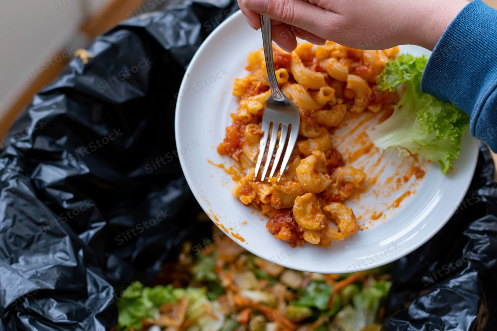 Hand scraping leftover tomato-sauced macaroni pasta off a plate into a black trash bag