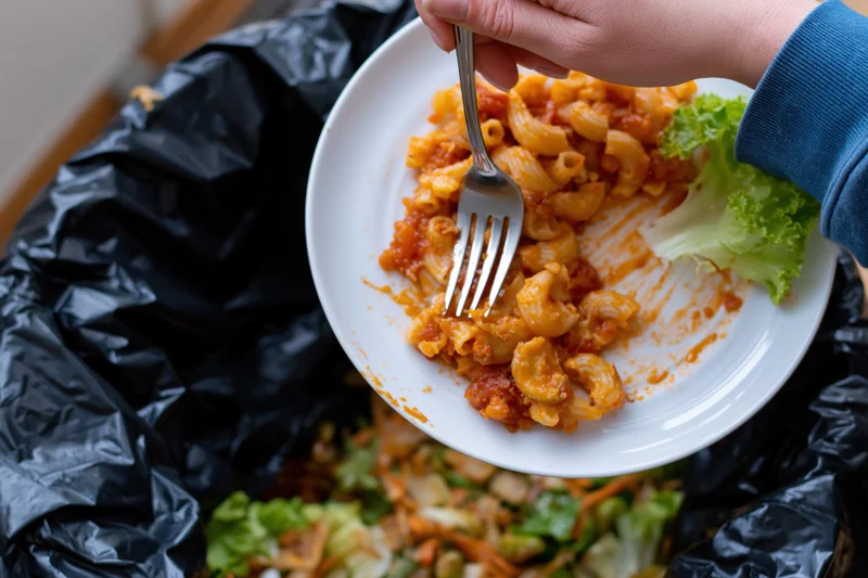 Hand scraping leftover tomato-sauced macaroni pasta off a plate into a black trash bag