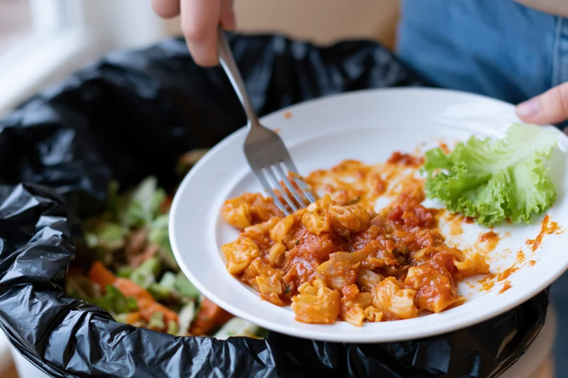 Person scraping leftover tomato-sauced macaroni pasta off a plate into a trash bin