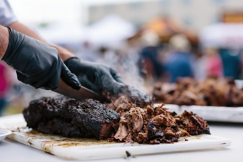 Pitmaster slicing and pulling smoked pork brisket on a cutting board at an outdoor food festival