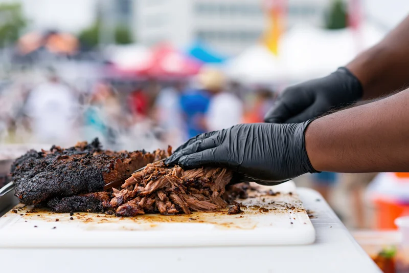 Pitmaster slicing and pulling smoked pork brisket on a white cutting board at an outdoor food festival