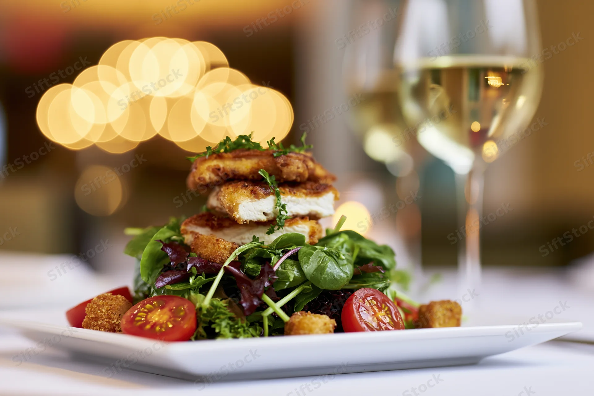 Plated salad with crispy chicken and mixed greens served with white wine on a restaurant table