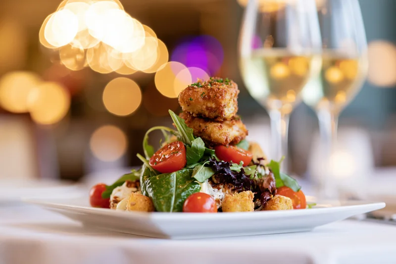 Plated salad with crispy chicken and mixed greens on a restaurant table with wine glasses and bokeh lights in the background