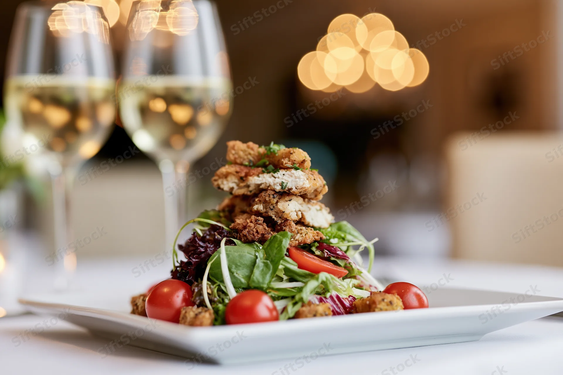 Plated salad with crispy chicken and mixed greens on a restaurant table with wine glasses and warm bokeh lighting in the background