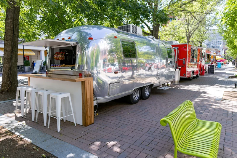 Polished silver airstream food truck with serving counter and stools on a brick plaza under trees
