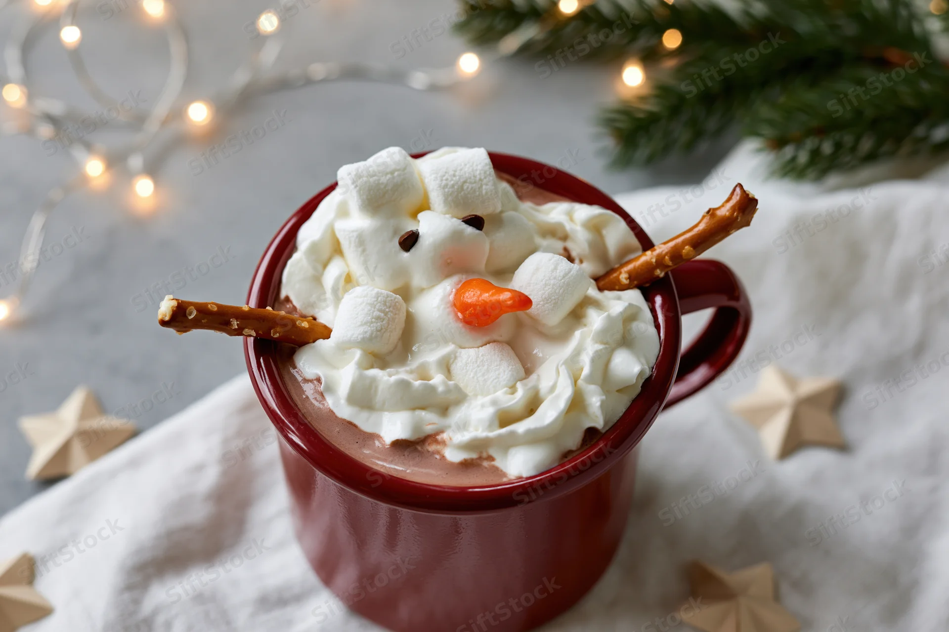 Red enamel mug filled with hot cocoa and whipped cream shaped as a snowman on a cloth with holiday lights and pine branches