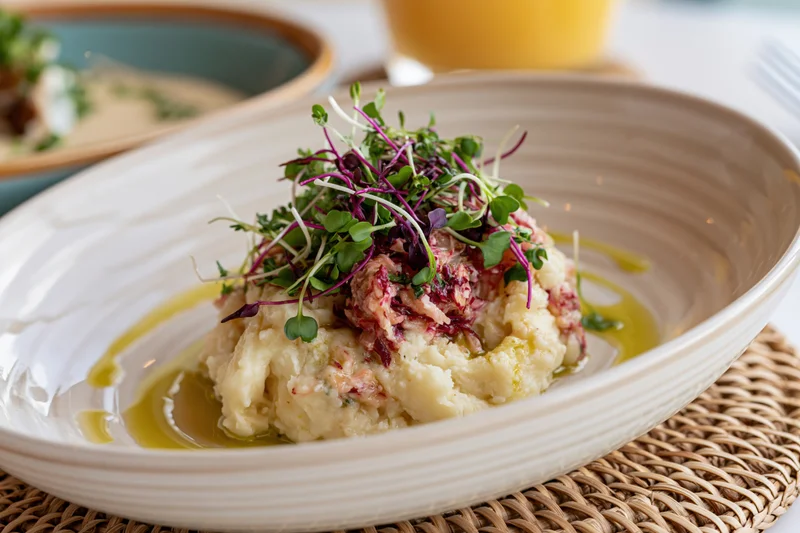 Mashed potato dish topped with microgreens and olive oil in a ceramic bowl on a woven placemat