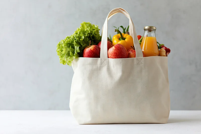Cream canvas tote bag filled with fresh produce and juice on a white surface