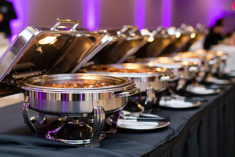 Row of stainless steel chafing dishes with open lids on a buffet table at an event