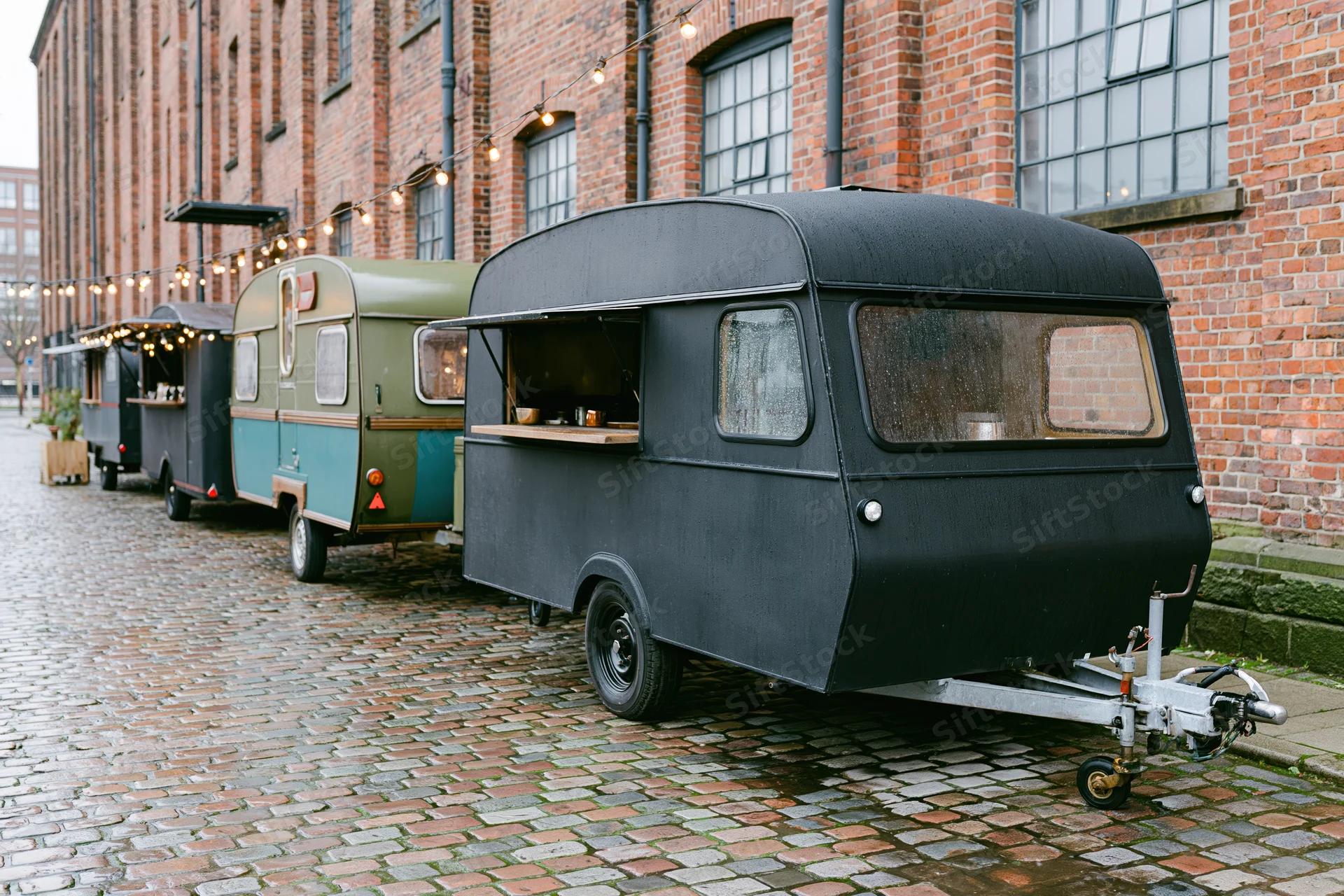 Vintage food trailers parked in a row on wet cobblestone street beside brick building