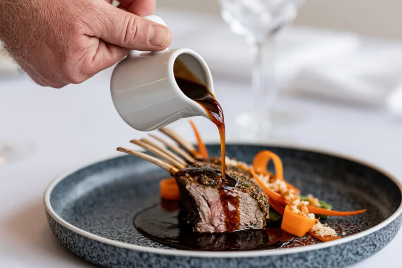 Hand pouring dark jus over a rack of lamb on a dark plate in a fine dining setting