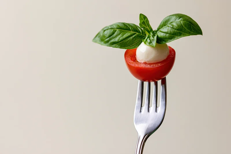 Fork holding halved cherry tomato with mozzarella and basil leaves against neutral background