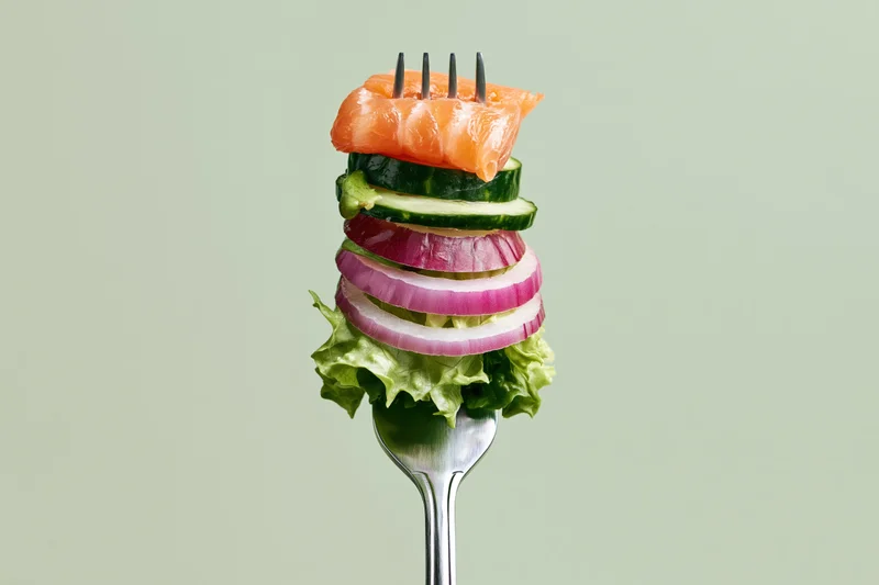 Fork holding stacked salmon, cucumber, red onion and lettuce against plain background