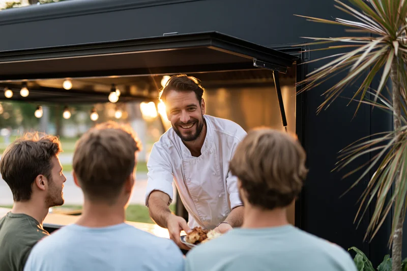 Smiling male chef serving food from a dark food truck window to customers outside