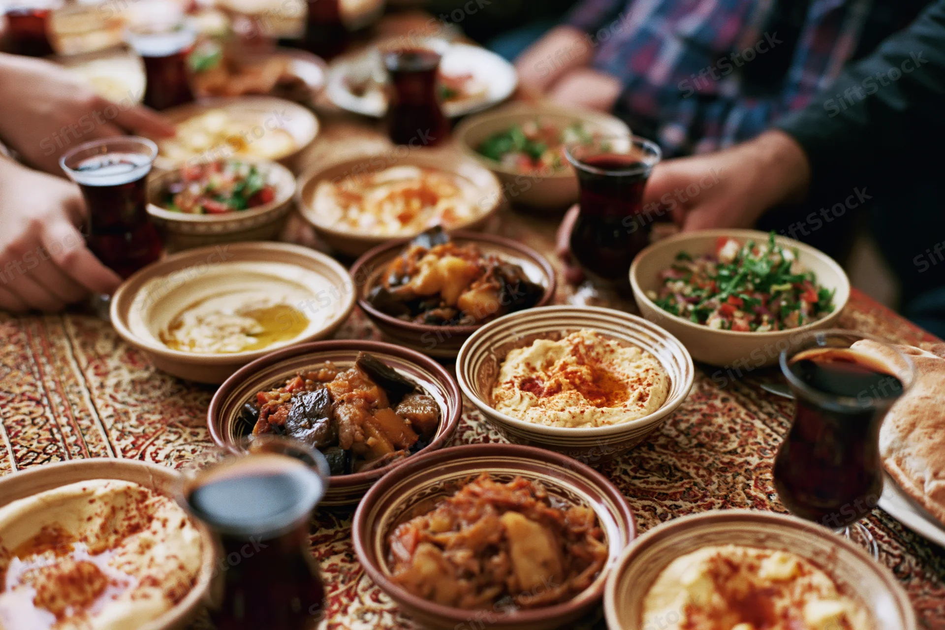 Middle eastern mezze dishes arranged on a patterned tablecloth with hands reaching for food and tea glasses around