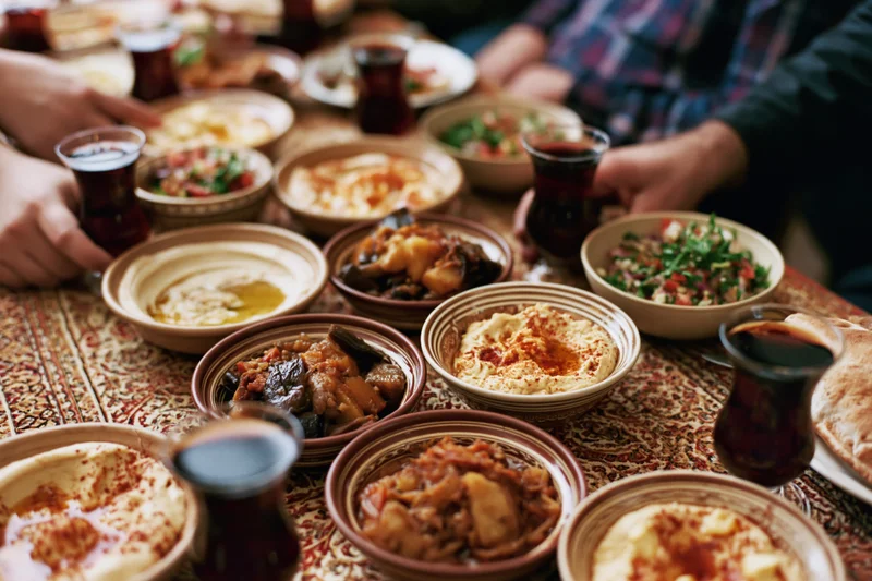 Middle eastern mezze dishes arranged on a patterned tablecloth with hands reaching for food and tea glasses around