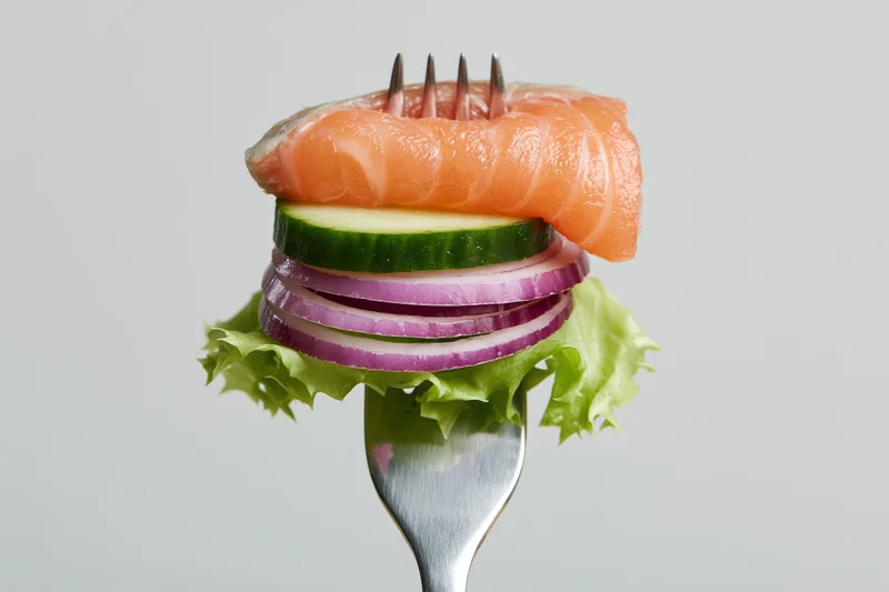 Fork holding stacked salmon, cucumber, red onion, and lettuce against white background