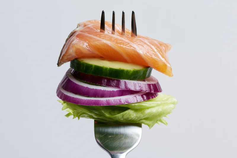 Fork holding stacked salmon, cucumber, red onion, and lettuce against white background