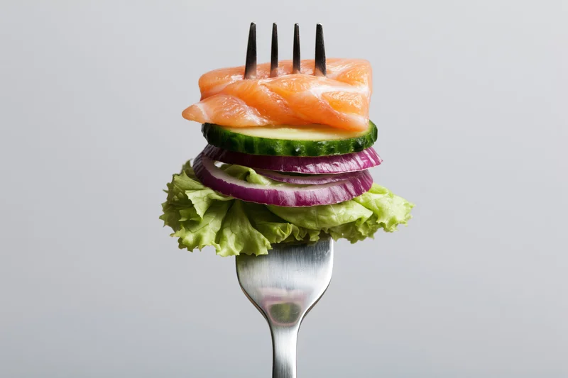 Fork holding stacked salmon, cucumber, red onion, and lettuce against white background