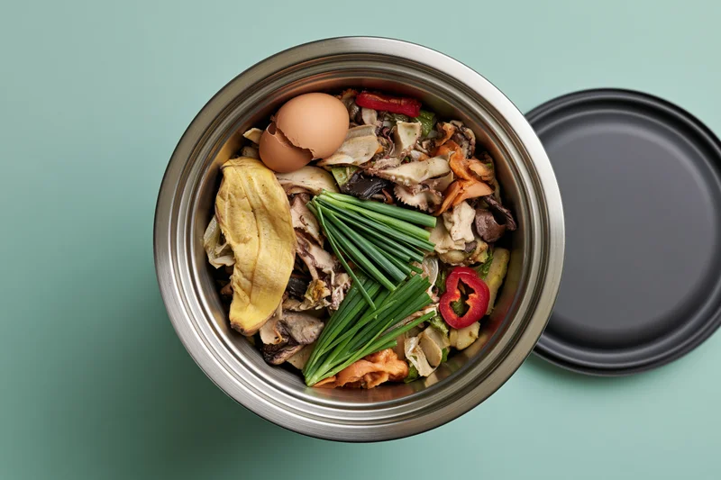 Stainless steel compost bin filled with mixed food scraps on a teal surface