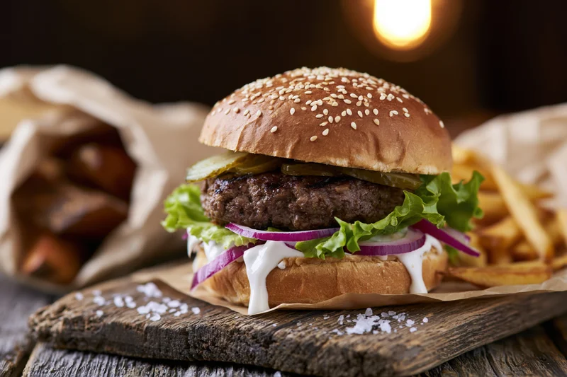Thick beef burger with lettuce, onion, pickle and sauce on a sesame brioche bun served with fries on a wooden board