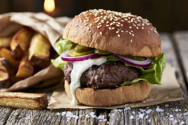 Thick beef burger with sesame brioche bun, lettuce, onion, pickle, and white sauce, served with fries on rustic wooden table