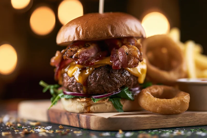 Thick beef burger with crispy bacon and melted cheddar on a wooden board with onion rings and fries beside it