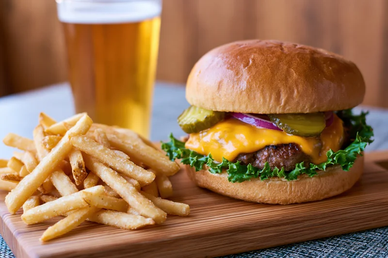 Cheeseburger with melted cheddar, pickles, red onion and lettuce on a wooden board beside fries and a glass of beer