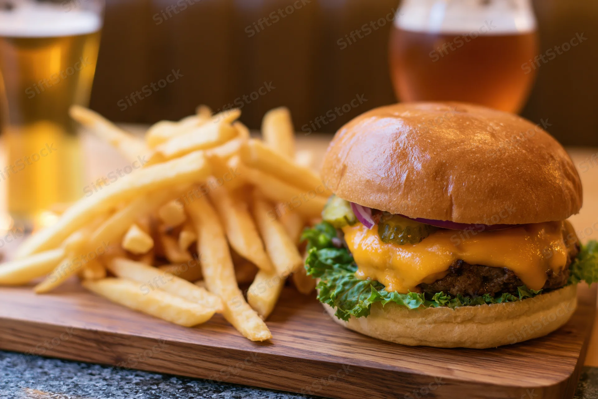 Cheeseburger with melted cheddar, pickles, lettuce and red onion served with french fries on a wooden cutting board beside two glasses of beer