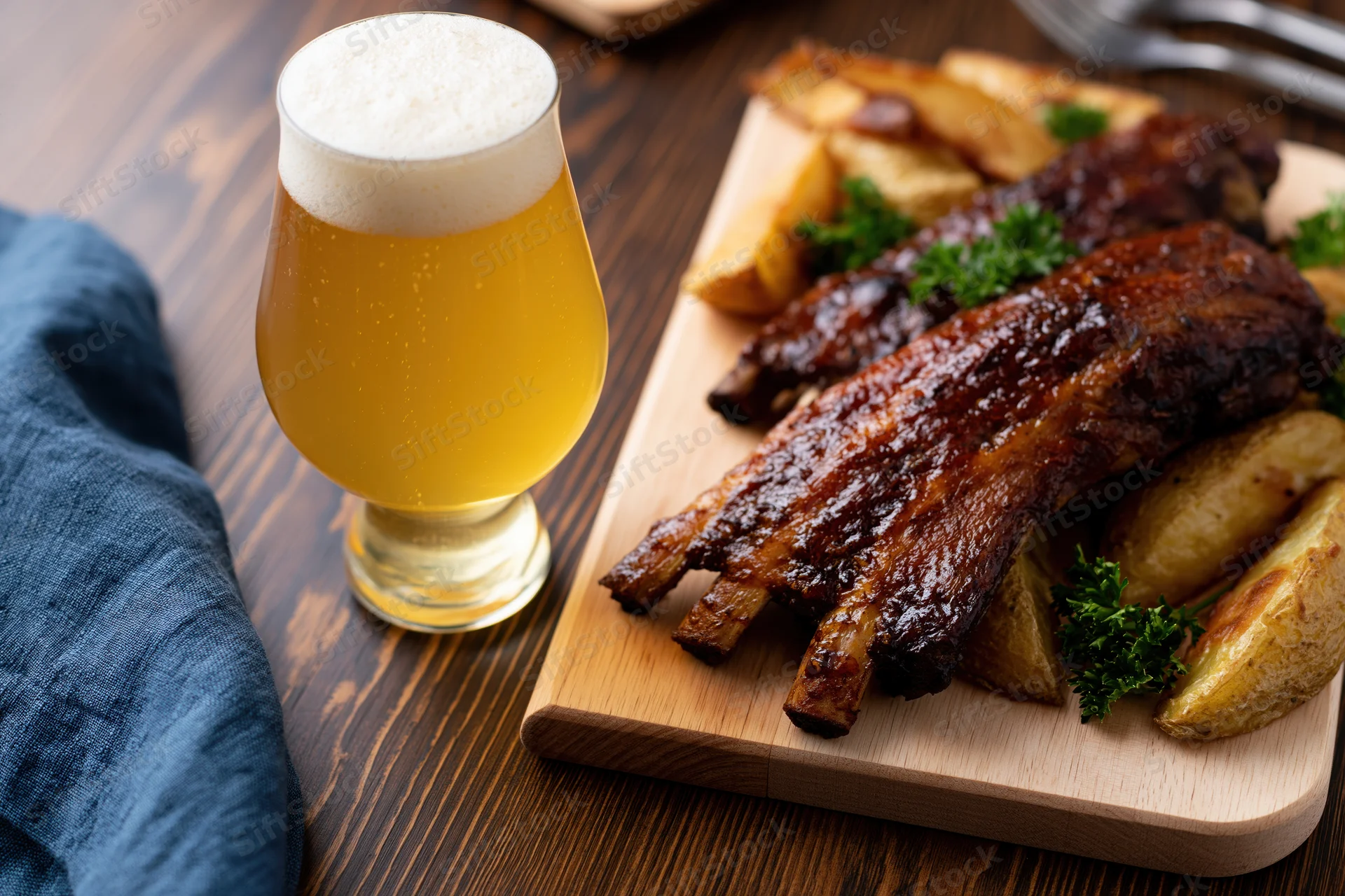 Tulip glass of pale lager with frothy head beside glazed pork ribs and potato wedges on wooden board