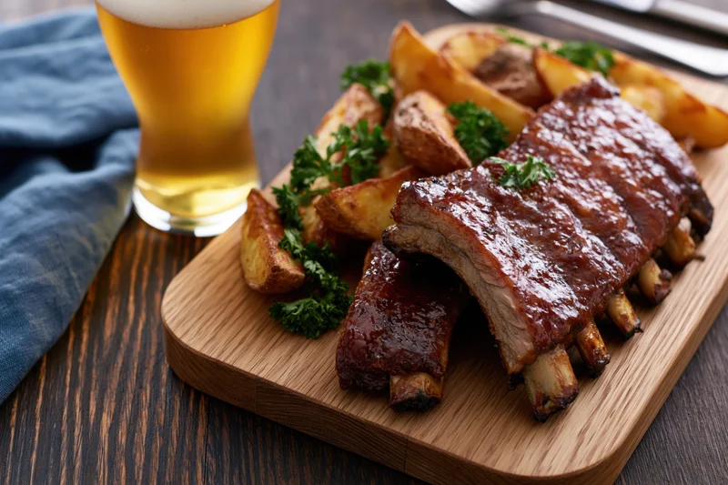 Glazed pork ribs with fries and parsley on a wooden cutting board beside a tulip glass of pale lager