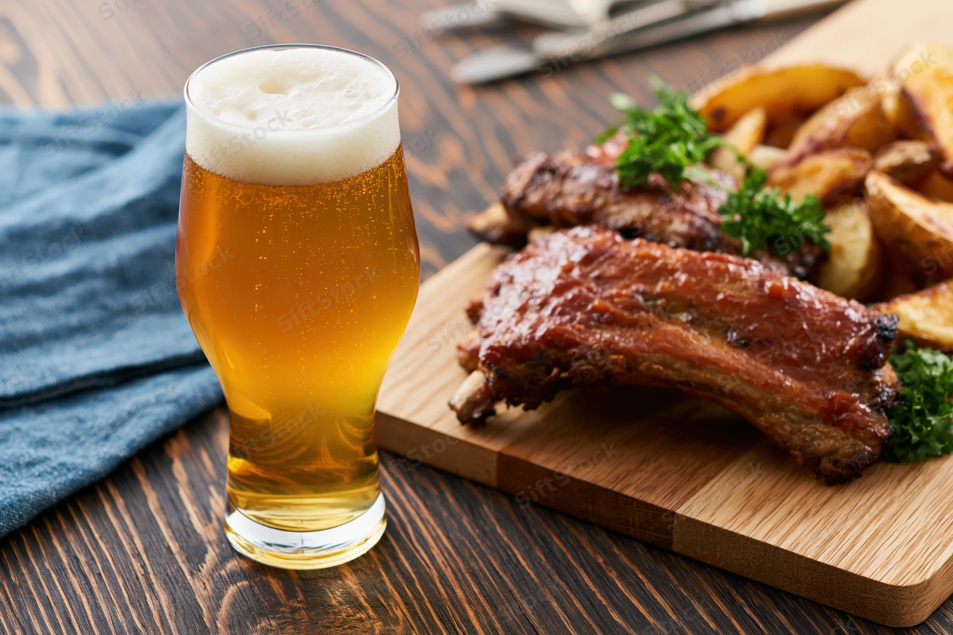 Tulip glass of pale lager with frothy head beside grilled ribs and potato wedges on wooden board
