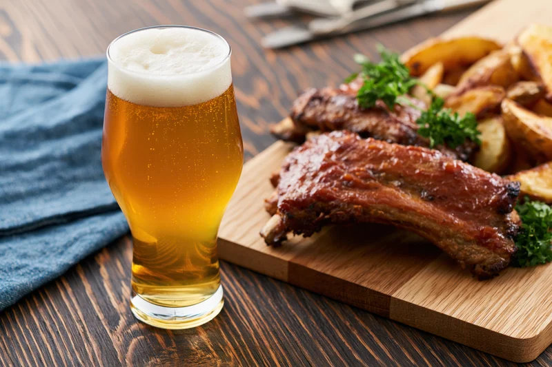 Tulip glass of pale lager with frothy head beside grilled ribs and potato wedges on wooden board