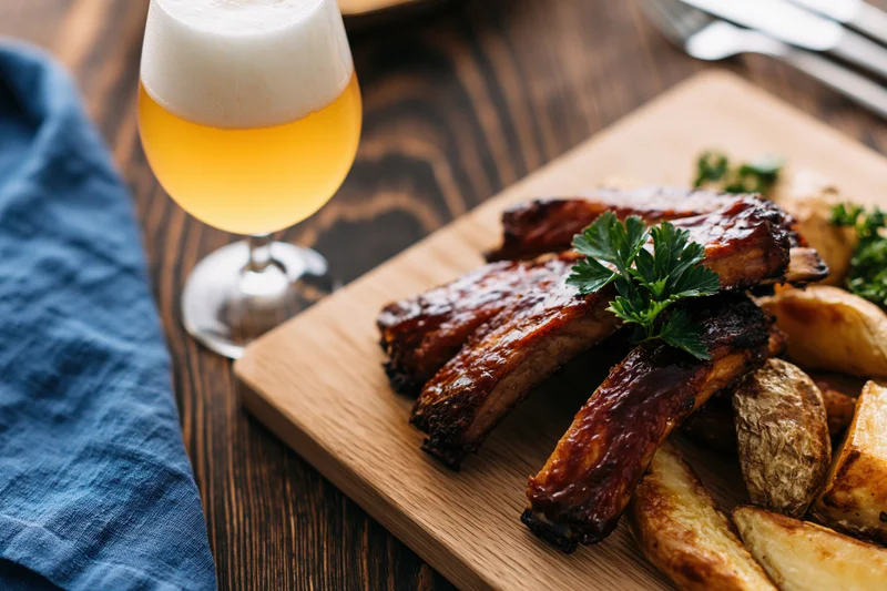 Glazed pork ribs with roasted potato wedges on a wooden cutting board beside a tulip glass of pale lager