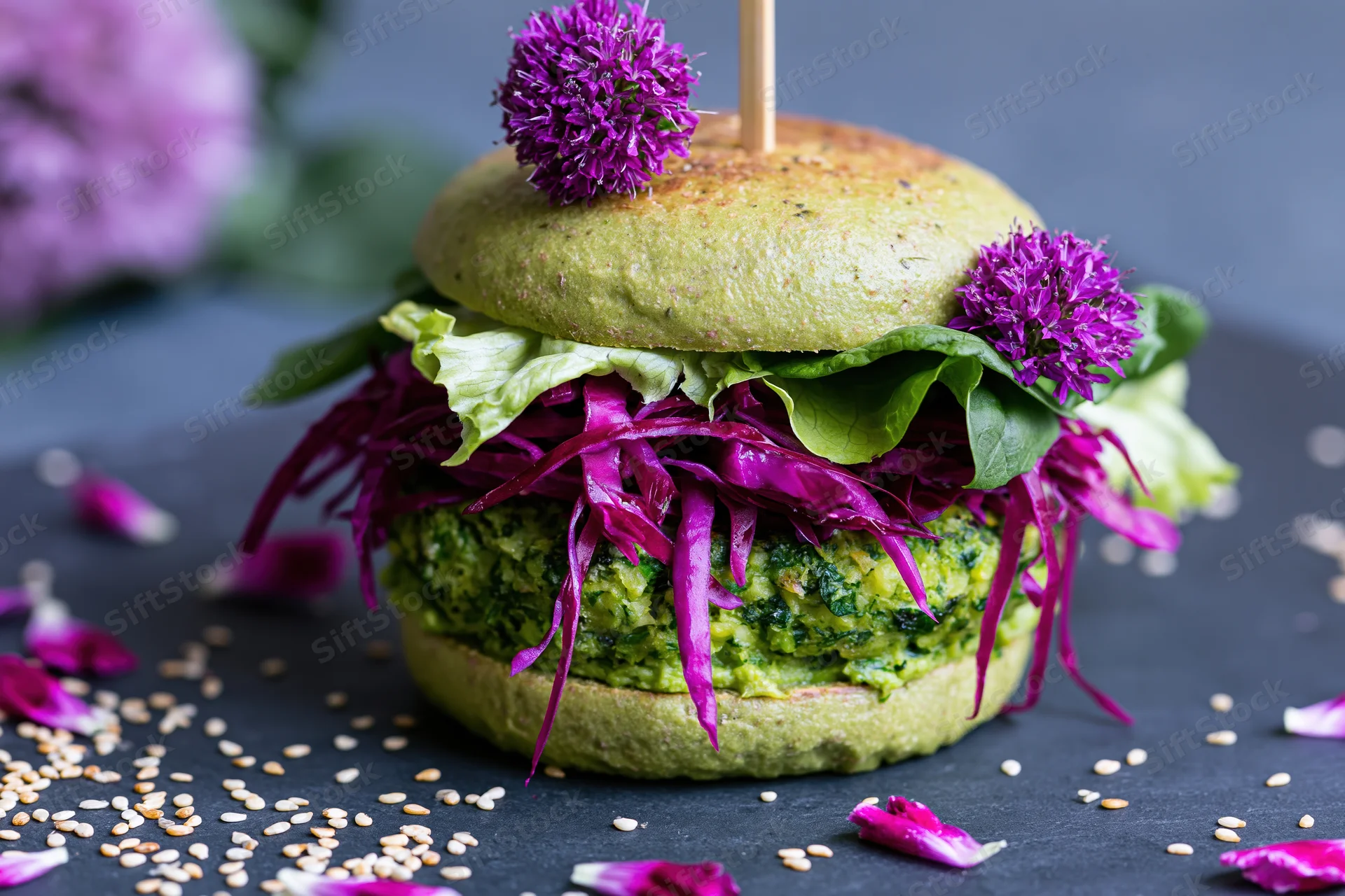 Vegan avocado bun burger with green herb patty and purple cabbage garnished with edible flowers on dark surface