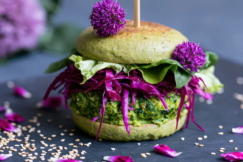 Vegan avocado bun burger with green herb patty and purple cabbage garnished with edible flowers on dark surface